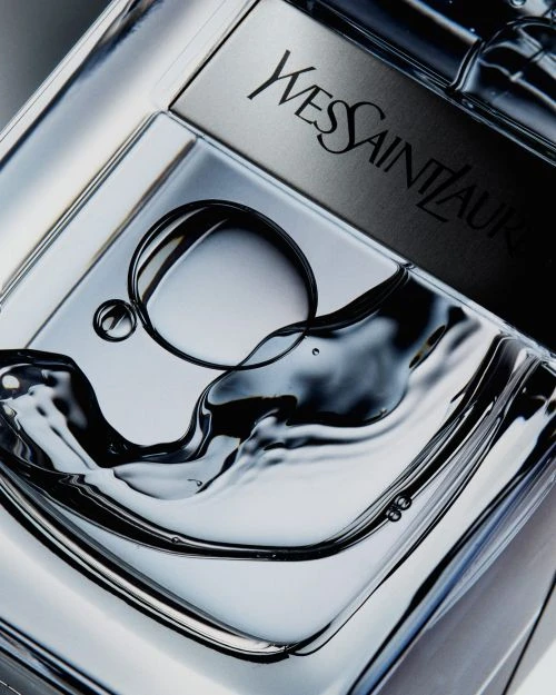 Close-up studio shot of a clear glass perfume bottle with Yves Saint Laurent branding, featuring liquid movement and sharp lighting.