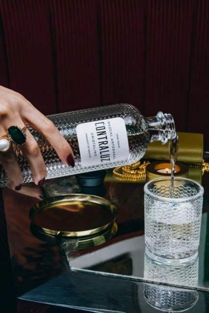 Pouring Contraluz Cristalino mezcal into a textured glass on a mirror, accented by gold accessories and a manicured hand.