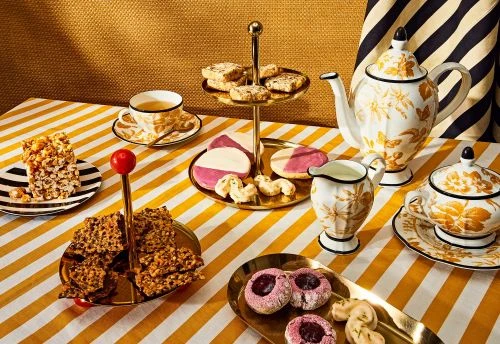 Overhead shot of an assortment of cookies, tea, and desserts arranged on a striped yellow and white tablecloth, presented in an indoor setting.