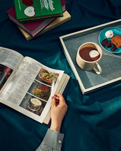 Open cookbook with food photos and stack of cookbooks next to a cup of hot chocolate with marshmallows on a tray.