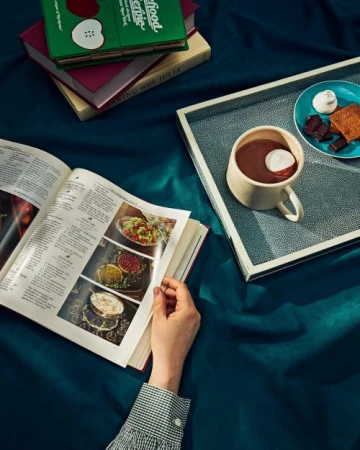 Open cookbook with food photos and stack of cookbooks next to a cup of hot chocolate with marshmallows on a tray.