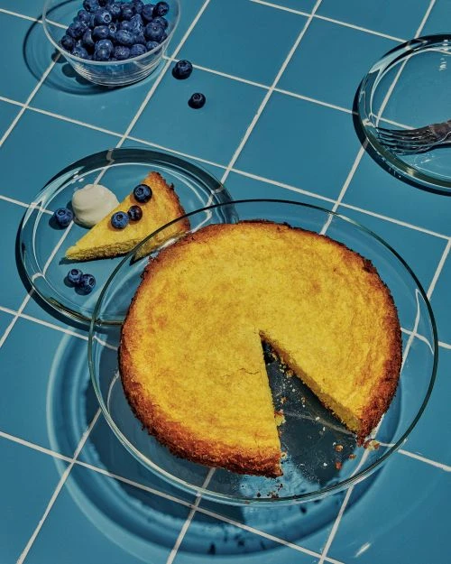 Overhead shot of a lemon cake with a slice cut out, topped with blueberries, next to a bowl of blueberries, on a blue tile surface.