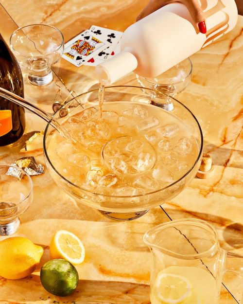 A punch bowl with ice and fruit slices filled with a light amber-colored drink being poured from a white bottle, with several glasses.