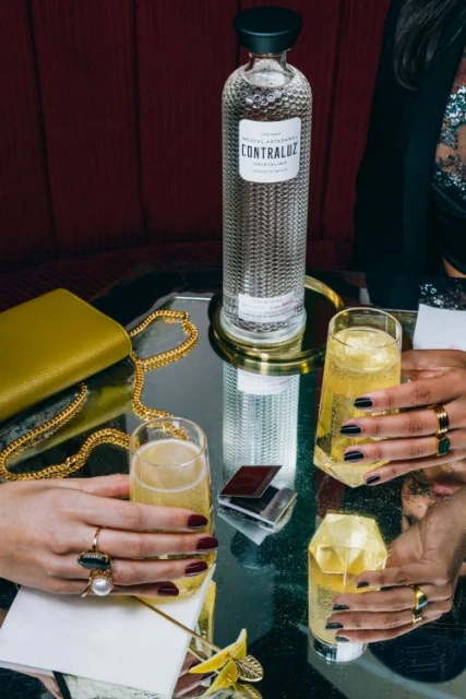 Elevated view of a Contraluz Cristalino Mezcal bottle, champagne flutes with cocktails, and women's hands holding the flutes.