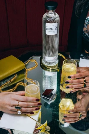 Elevated view of a Contraluz Cristalino Mezcal bottle, champagne flutes with cocktails, and women's hands holding the flutes.