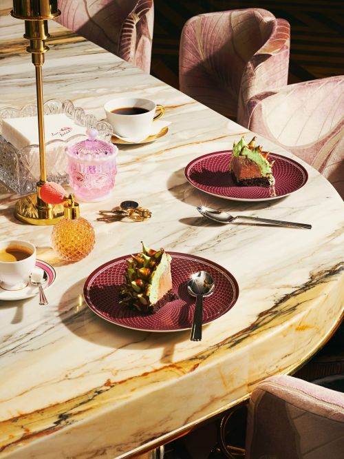 Overhead shot of a table set with slices of cake, cups of coffee, perfume bottles and keys, accented by pink chairs and elegant lighting.