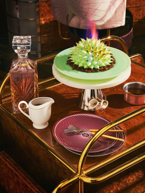 Cake on a cake stand with a decanter, plates, and creamer on a bar cart, showcasing gourmet food presentation with golden accents.