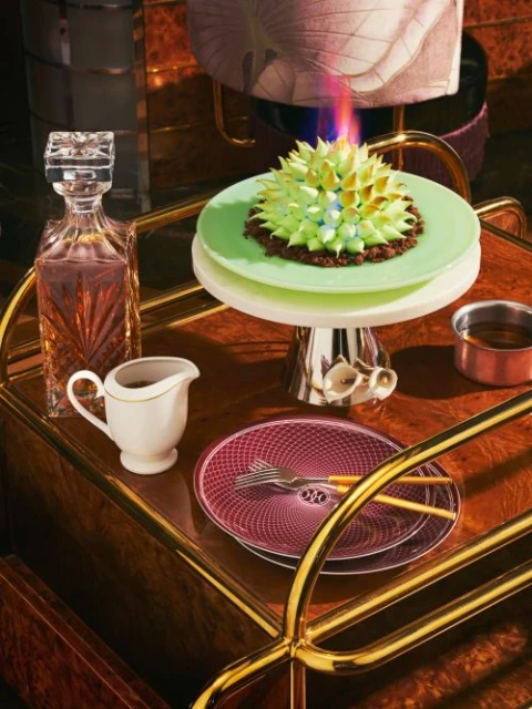 Cake on a cake stand with a decanter, plates, and creamer on a bar cart, showcasing gourmet food presentation with golden accents.