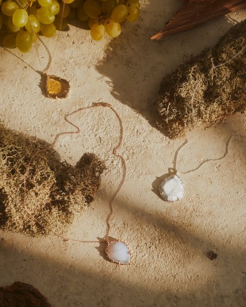 Three necklaces with gemstone pendants are displayed on a textured surface with grapes and moss, creating an earthy, luxurious feel.