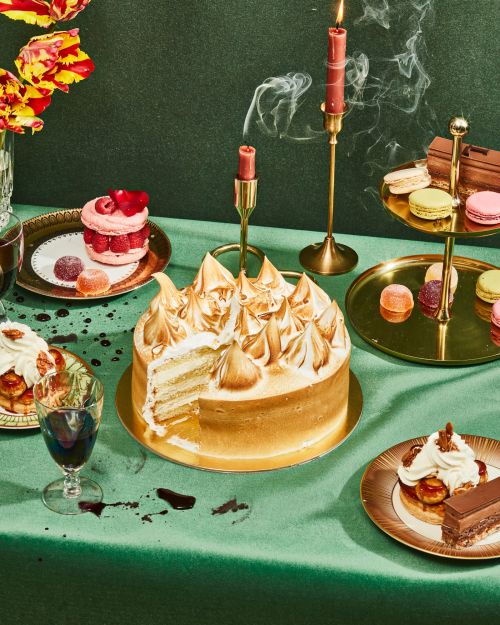Elegant dessert display with a sliced meringue cake, macarons, and pastries arranged on a green tablecloth with burning candles.