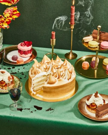 Elegant dessert display with a sliced meringue cake, macarons, and pastries arranged on a green tablecloth with burning candles.
