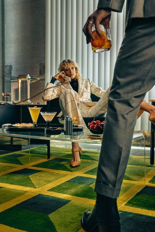Woman in suit talking on telephone while man in suit holds cocktail. Retro style indoor scene with cocktails and geometric rug.