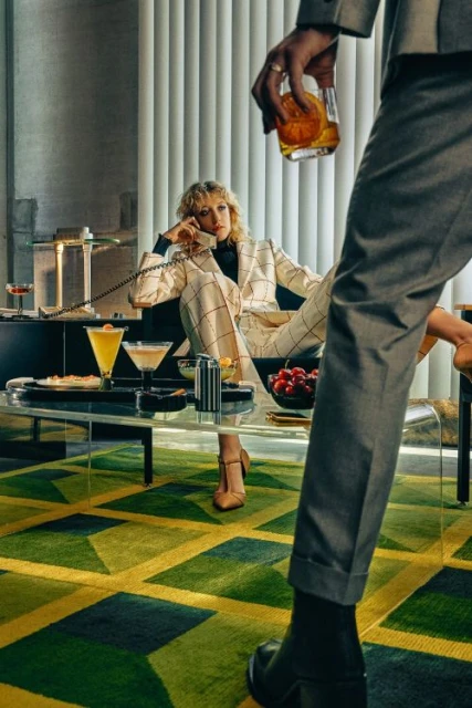 Woman in suit talking on telephone while man in suit holds cocktail. Retro style indoor scene with cocktails and geometric rug.
