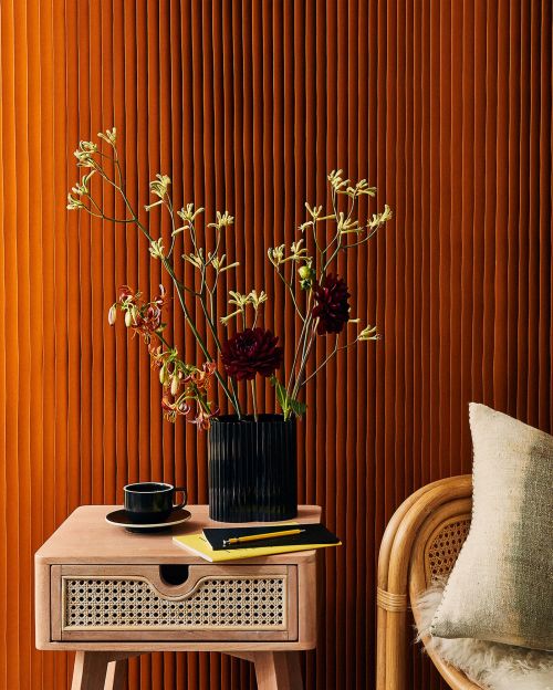 Interior still life featuring an end table with a vase of flowers, a cup, and a book against a textured brown background wall.