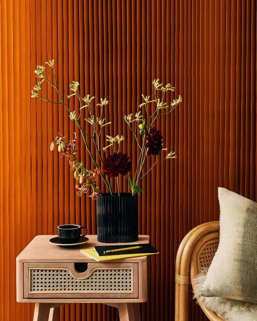Interior still life featuring an end table with a vase of flowers, a cup, and a book against a textured brown background wall.