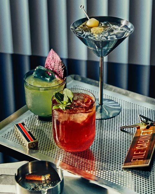 Three colorful cocktails on a silver tray with an ashtray, lighter, hotel key and ice. Blue blinds provide a moody backdrop.