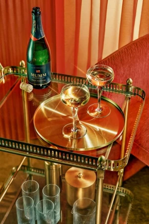 Close-up of a Mumm champagne bottle and two coupe glasses filled with sparkling wine on a gold bar cart against an orange background.