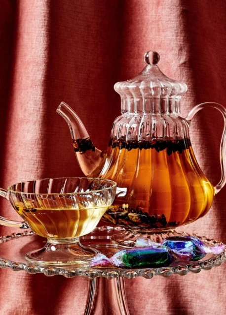 Elegant glass teapot and teacup on a cake stand filled with tea, accompanied by candies and set against a red draped fabric backdrop.