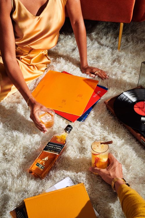 Two cocktails next to a whiskey bottle, surrounded by records on a white fluffy rug, with two people relaxing at home.
