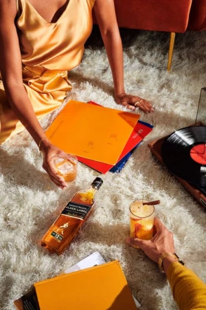 Two cocktails next to a whiskey bottle, surrounded by records on a white fluffy rug, with two people relaxing at home.