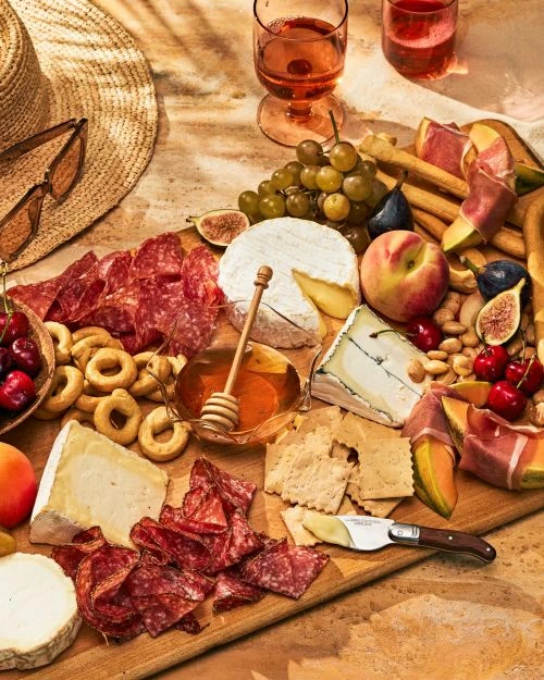 Overhead shot of a wooden charcuterie board featuring cheese, salami, fruits, nuts, crackers, honey, and glasses of pink wine.