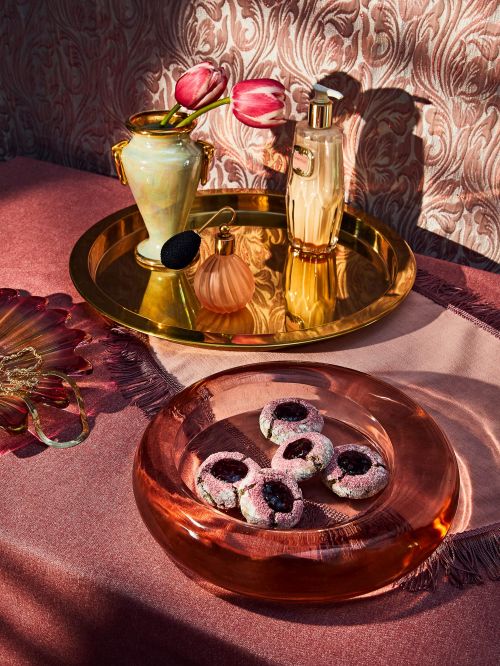 Still life featuring a bowl of cookies, perfume bottles, tulips in a vase, and a golden tray, set against a patterned backdrop.