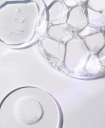 Close-up macro shot of transparent cosmetic gel, soapy foam bubbles, and white powder on a white surface.