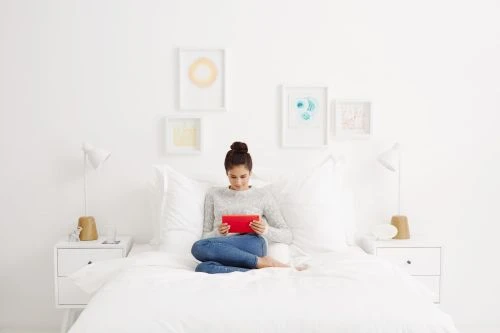 A young adult woman with a hair bun sits cross-legged on a white bed, holding a red tablet. Minimalist decor and bright daylight.