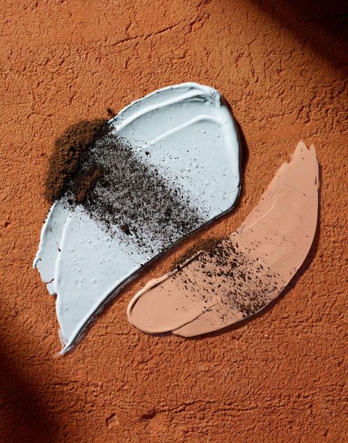 Close-up of light blue and peach-colored cosmetic cream swatches sprinkled with brown granules on a textured terracotta surface.