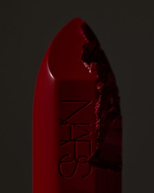 Close-up of a red NARS lipstick with a broken tip on a dark background, showcasing cosmetic textures and product detail in studio lighting.