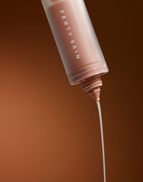Close-up shot of a Fenty Skin product bottle with a stream of the light-colored skincare product being dispensed against a brown background.