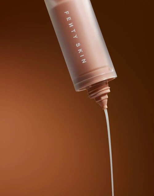 Close-up shot of a Fenty Skin product bottle with a stream of the light-colored skincare product being dispensed against a brown background.