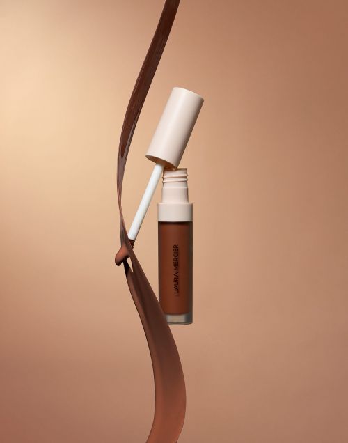 Close-up studio shot of Laura Mercier concealer bottle and applicator wand with splash of product on a tan background.
