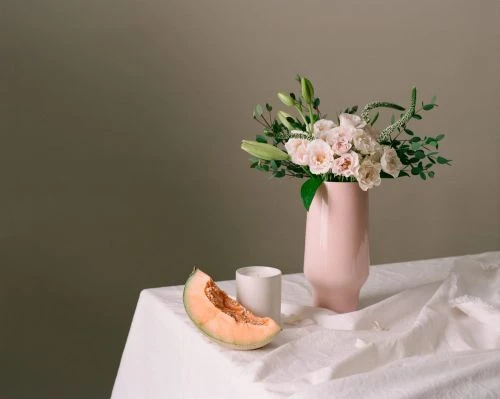Floral arrangement in a pink vase with a melon slice and a white mug on a table against a neutral grey background. Still life.