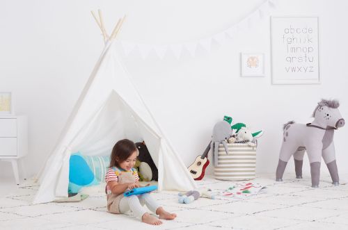 Child plays with a tablet inside a white teepee surrounded by stuffed animals, with neutral carpet and walls in a bright, indoor setting.