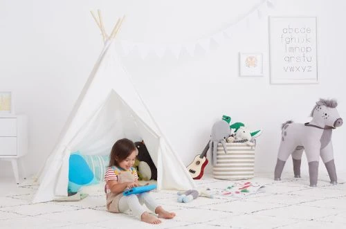 Child plays with a tablet inside a white teepee surrounded by stuffed animals, with neutral carpet and walls in a bright, indoor setting.