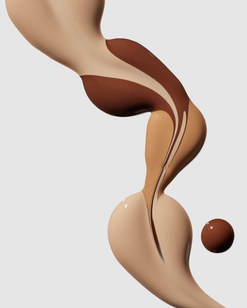 Swirls of varying shades of liquid foundation, including light tan and dark brown, create an abstract design on a light gray background.
