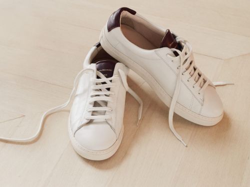 Pair of white leather sneakers with white laces and burgundy heel detail on a light wood floor, close-up fashion product shot.