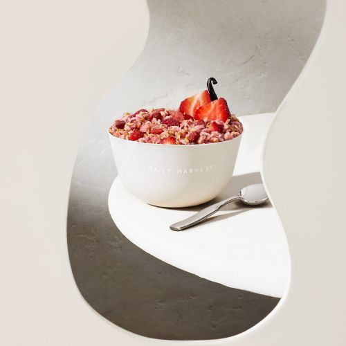 Bowl of granola topped with sliced strawberries and a vanilla bean on a white table with a spoon, shot through a geometric frame.