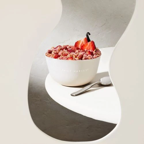 Bowl of granola topped with sliced strawberries and a vanilla bean on a white table with a spoon, shot through a geometric frame.