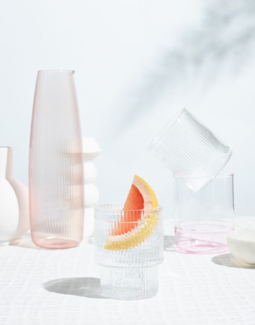Still life of assorted glass drinkware and a grapefruit slice, set on a white textured tablecloth with a white background.