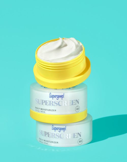 Stacked Supergoop! sunscreen containers in yellow and white on a bright blue background, close-up studio product shot for beauty and skincare.