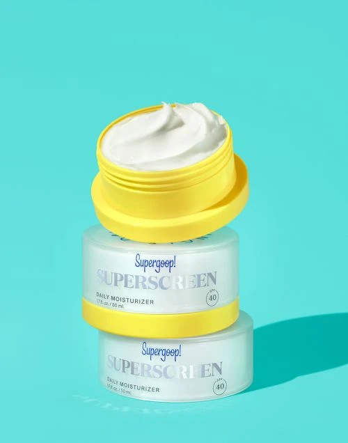 Stacked Supergoop! sunscreen containers in yellow and white on a bright blue background, close-up studio product shot for beauty and skincare.
