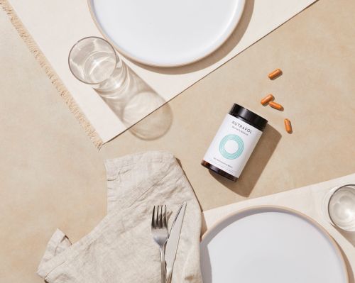 Top-down shot of Nutrafol supplement bottle with capsules, a drinking glass, plates, and cutlery arranged on a beige surface.