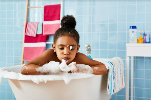 A young Black girl sitting in a white bathtub playing with soap bubbles in a blue tiled bathroom styled by Rebecca Donnelly.