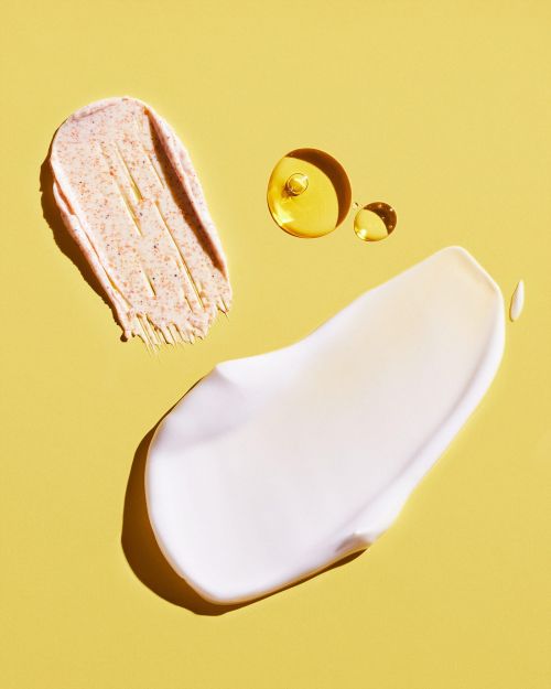 Three blobs of skincare products on a yellow background: a scrub, a white cream, and clear oil drops.
