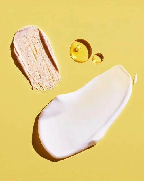 Three blobs of skincare products on a yellow background: a scrub, a white cream, and clear oil drops.