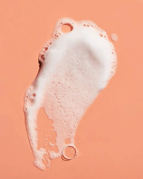 Close-up of a dollop of foamy white cleanser against a peach-colored surface. Skincare and personal care concept with bright, direct lighting.
