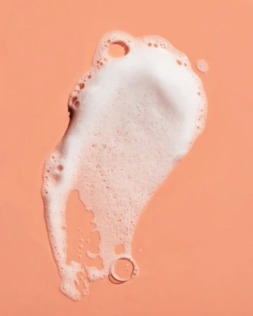 Close-up of a dollop of foamy white cleanser against a peach-colored surface. Skincare and personal care concept with bright, direct lighting.