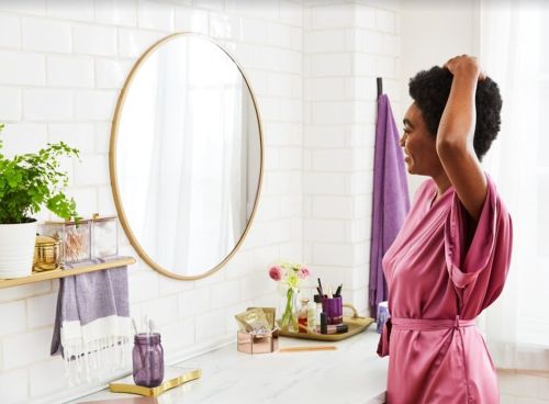 Woman in a satin robe fixes her hair in a bathroom with white subway tile and decor, captured in a bright and inviting scene.